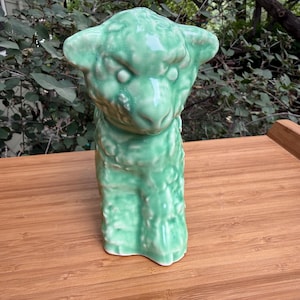 May include: A ceramic figurine of a lamb with a glossy, light green glaze. The sculpture features a textured surface representing the woolly coat of the animal, with defined ears, eyes, and a snout, standing in a forward-facing position.