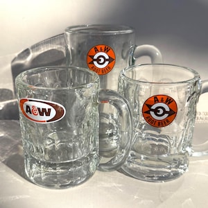 May include: Three clear glass A&W Root Beer mugs with handles. Each mug has a textured exterior and a circular logo with the A&W logo and the words "Root Beer". The mugs are empty and appear to be vintage.