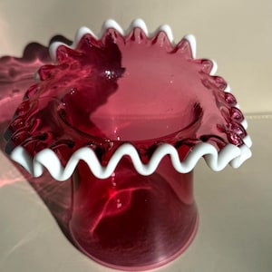 May include: A ruby red glass vase with a ruffled, white-edged rim. The vase has a cylindrical body and a flared top, with the white trim adding a decorative touch. The glass is transparent, allowing light to pass through.