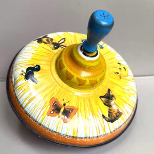 May include: Vintage metal spinning top toy with a yellow and orange design. The top features illustrations of a butterfly, kitten, chick, rabbit, and bird. It has a blue handle and a yellow and orange base.