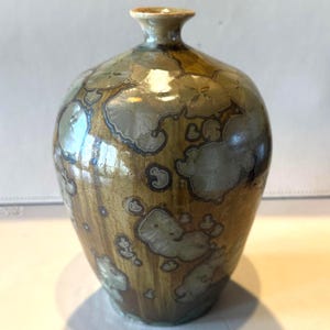 May include: A handcrafted ceramic vase with a unique crystalline glaze. The vase features a bulbous body and a narrow neck. The glaze has a mix of brown, green, and silver tones, creating a textured, organic appearance. The vase is approximately 10 inches tall.