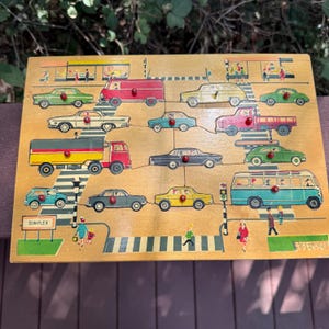 May include: A rectangular wooden peg puzzle featuring a colorful street scene with various vehicles including cars, a truck, and a bus. Each vehicle has a small red knob for handling. The board displays crosswalks, traffic lights, and the text SIMPLEX.