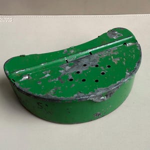 May include: A vintage, green metal container with a curved top and a series of small holes. The container has a worn, distressed appearance with visible signs of age and use. The bottom of the container has the number "5" written on it.