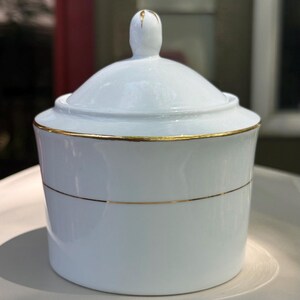 May include: White porcelain sugar bowl with a lid and gold trim. The bowl has a cylindrical shape with a gold band around the middle. The lid has a small, rounded handle with gold accents. The sugar bowl is a classic piece for serving sugar or other condiments.