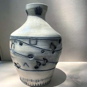 May include: A ceramic vase with a light gray, off-white glaze and a dark blue, incised musical staff pattern wrapping around the body. The design features carved musical notes, symbols, and a decorative border at the base with a narrow neck.