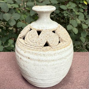 May include: A handcrafted, off-white ceramic vase with a textured surface and a unique design. The vase features a series of cut-out shapes around the top, adding a decorative element. The vase is approximately 8 inches tall, with a rounded body and a narrow neck.