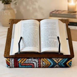 May include: A wooden book stand with a dark brown finish and two black metal page holders, featuring a base cushion decorated with a colorful geometric embroidered pattern in shades of red, yellow, blue, and black, holding an open hardcover book.
