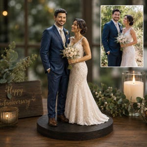 Könnte beinhalten: Ein individueller Tortenaufsatz für Hochzeiten mit einem Miniaturpaar auf einem dunklen runden Sockel. Die Figur trägt einen marineblauen Anzug und die andere ein weißes Spitzenkleid und hält einen Strauß heller Blumen, passend zum Foto.