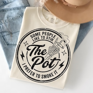 Puede incluir: Camiseta color crema con un gráfico negro. El gráfico presenta una mano de esqueleto sosteniendo un cigarrillo encendido, con el texto: "Some people like to stir the pot, I prefer to smoke it."