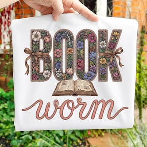 May include: White tote bag with the words "BOOK WORM" in a floral design. The word "BOOK" is in large, ornate letters filled with colorful flowers. A brown ribbon is tied around the "B" and the "K". An open book is below the word "BOOK".