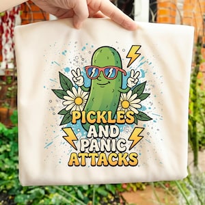 May include: A cream-colored tote bag featuring a cartoon pickle wearing sunglasses and making a peace sign. The text "PICKLES AND PANIC ATTACKS" is displayed in a colorful, playful font, with lightning bolt graphics.