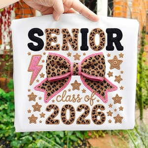 Puede incluir: Camiseta blanca con la palabra "SENIOR" en negro y estampado de leopardo, y "class of 2026". El diseño incluye un lazo con estampado de leopardo, detalles en rosa, un rayo rosa y estrellas.