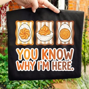 Peut inclure: T-shirt noir avec un motif de trois œufs farcis et le texte "YOU KNOW WHY I'M HERE." Les œufs sont de style dessin animé avec des accents orange et jaune. Le t-shirt est tenu par une main.