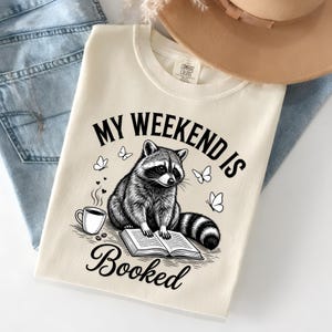 May include: Cream-coloured sweatshirt featuring a raccoon illustration reading a book, with butterflies and a coffee cup. The text "MY WEEKEND IS Booked" is printed above and below the raccoon graphic. The sweatshirt is folded on top of blue jeans.