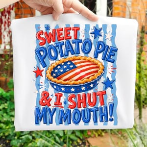 Può includere: T-shirt bianca con una grafica di torta di patate dolci decorata con la bandiera americana. Il testo recita "Sweet Potato Pie & I Shut My Mouth!" in rosso e blu, con stelle e strisce.