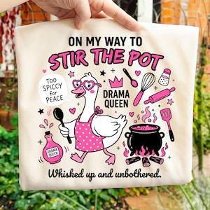 Puede incluir: Bolso tote de color crema con una ilustración caprichosa. Presenta un ganso de dibujos animados con gafas en forma de corazón y un delantal, sosteniendo una cuchara. El texto incluye "ON MY WAY TO STIR THE POT" y "DRAMA QUEEN".