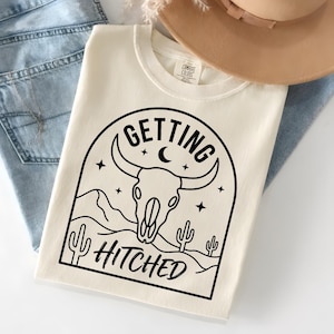 Könnte beinhalten: Cremefarbenes T-Shirt mit schwarzer Grafik eines Bullenschädels, Kakteen und den Worten "Getting Hitched". Das Design wird von einem Bogen mit Sternen und einem Halbmond umrahmt. Ein beigefarbener Hut und blaue Jeans sind im Hintergrund.