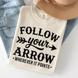 Może przedstawiać: Kremowa koszulka z czarnym napisem "FOLLOW your ARROW - WHEREVER IT POINTS". Koszulka jest złożona i leży na niebieskich dżinsach, z brązowym kapeluszem w tle. Materiał jest miękki.