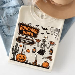 Peut inclure: Sweat-shirt crème avec un motif sur le thème d'Halloween. Le motif représente un squelette en patins à roulettes, tenant des assiettes de nourriture, et le texte "Boneyard Diner Serving Spirits Since 1666." Comprend des citrouilles, des chauves-souris, une toile d'araignée et un cactus.