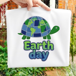 Puede incluir: Bolso tote blanco con un diseño de tortuga de dibujos animados. El caparazón de la tortuga es un mapa del mundo en verde y azul. Debajo de la tortuga están las palabras "Earth day" en verde y azul.