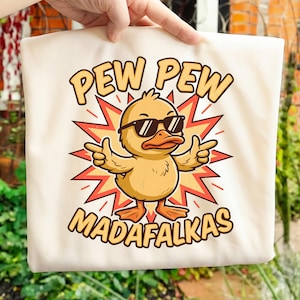May include: Cream-colored tote bag featuring a cartoon duck wearing sunglasses and making a hand gesture. The duck is set against a red and yellow starburst design. Text on the bag reads "PEW PEW MADAFALKAS" in a bold, playful font.
