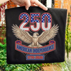 Puede incluir: Camiseta negra con un diseño patriótico. El diseño incluye el número "250" en rojo y azul, un águila con las alas extendidas y el texto "AMERICAN INDEPENDENCE 1776-2026" en una pancarta.