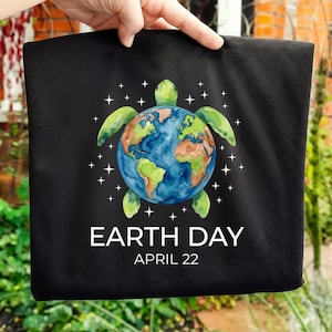 Puede incluir: Bolso tote negro con una ilustración de acuarela de una tortuga con la Tierra como caparazón. El diseño incluye el texto "EARTH DAY APRIL 22" en blanco. La tortuga es verde y la Tierra es azul y marrón.