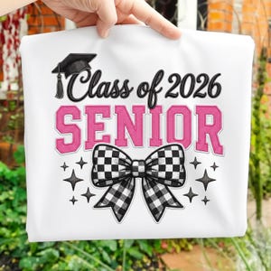 Op de afbeelding: Wit T-shirt met de tekst "Class of 2026 SENIOR" in zwart en roze. Voorzien van een afstudeerhoed en een zwart-wit geblokte strik. Inclusief sterren.