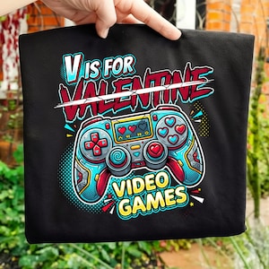 Könnte beinhalten: Schwarzes T-Shirt mit einer farbenfrohen Grafik eines Videospiel-Controllers mit herzförmigen Knöpfen. Der Text "V IS FOR VALENTINE VIDEO GAMES" ist oberhalb und unterhalb des Controllers in einer verspielten Schriftart dargestellt, ideal für Valentinstag.