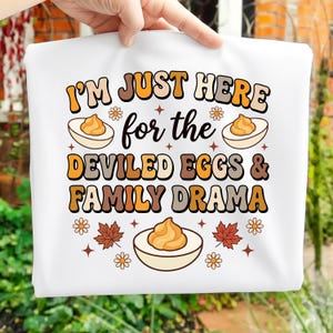 May include: White tote bag featuring the text "I'M JUST HERE for the DEVILED EGGS & FAMILY DRAMA" in orange, brown, and cream. The design includes illustrations of devilled eggs, flowers, and leaves.