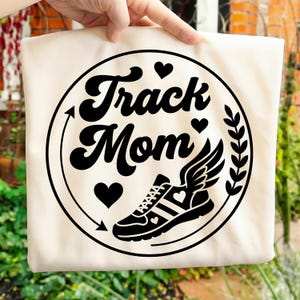 Op de afbeelding: Crèmekleurige draagtas met een zwarte afbeelding. Het ontwerp bevat de woorden "Track Mom", een gevleugelde hardloopschoen, harten en een lauwerkrans, allemaal binnen een cirkel. De tas wordt door een hand omhoog gehouden.