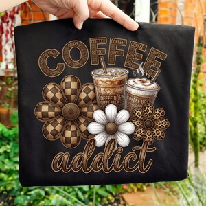 Può includere: T-shirt nero con un design a tema caffè. La parola "COFFEE" è visualizzata impilata, con "addict" sotto. Il design include tazze di caffè e grafiche floreali. Il testo "COFFEE BREW" è anche visibile.