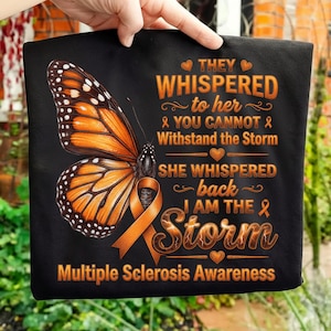 May include: Black t-shirt featuring a large orange and black butterfly graphic. The shirt includes text that reads "THEY WHISPERED to her & YOU CANNOT Withstand the Storm SHE WHISPERED back I AM THE Storm Multiple Sclerosis Awareness."