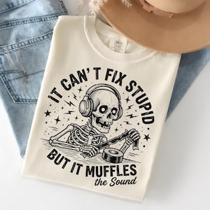 May include: Cream-colored t-shirt with a black and white graphic of a skeleton wearing headphones and holding tape. The text reads "IT CAN'T FIX STUPID BUT IT MUFFLES the SOUND". The shirt is folded on top of blue jeans.