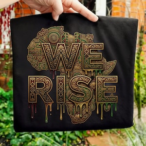 Puede incluir: Camiseta negra con un mapa detallado de África en verde, dorado y marrón. Las palabras "WE RISE" se muestran en una fuente goteante y texturizada. El diseño tiene un tema cultural.