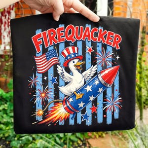 Könnte beinhalten: Schwarzes T-Shirt mit einem patriotischen Design. Es zeigt eine Ente mit einem Uncle Sam-Hut, die eine amerikanische Flagge hält und auf einer Rakete reitet. Das Wort "FIREQUACKER" steht in roten Buchstaben über der Grafik. Feuerwerk und Sterne vervollständigen das Design.
