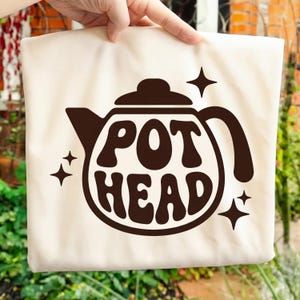 Puede incluir: Bolso tote color crema con un diseño de tetera marrón y las palabras "POT HEAD" en una fuente negrita y redondeada. Cuatro estrellas rodean el gráfico de la tetera. El bolso está sostenido por una mano.