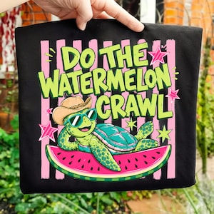Peut inclure: T-shirt noir avec l'inscription "Do the Watermelon Crawl" en vert et rose. Le graphisme représente une tortue de dessin animé portant des lunettes de soleil et un chapeau de cow-boy, allongée sur une tranche de pastèque.