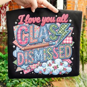 May include: Black t-shirt with the text "I love you all CLASS DISMISSED" in a colorful, patchwork design. The design includes a pencil, lightning bolt, and floral accents, perfect for teachers.