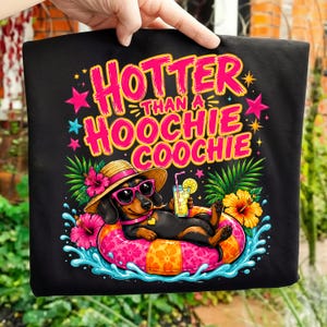 May include: Black t-shirt with a colorful graphic of a dachshund wearing sunglasses and a straw hat, relaxing on an inflatable ring in water. Text reads "Hotter Than A Hoochie Coochie."