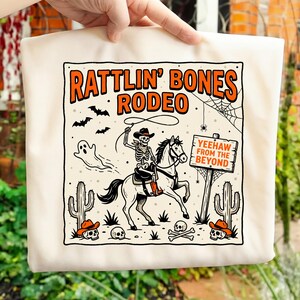 Könnte beinhalten: Beige Tragetasche mit einem Halloween-Design. Das Design zeigt den Text "Rattlin' Bones Rodeo", ein Skelett auf einem Pferd, Fledermäuse, einen Geist, Kakteen und Totenköpfe. Das Schild lautet "Yeehaw From The Beyond".