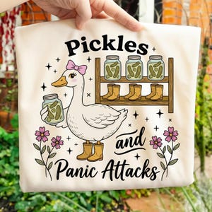 Può includere: Borsa tote color crema con un'oca dei cartoni animati con un fiocco rosa e un barattolo di sottaceti. La borsa ha le parole "Pickles and Panic Attacks" in nero, con uno scaffale di sottaceti e stivali.