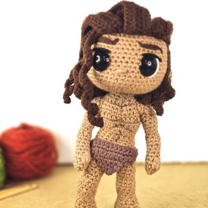 May include: A handmade crochet doll with a tan complexion, brown hair, and large black eyes. The doll is wearing a brown loincloth. The doll's hair is styled in a curly fashion. The doll is standing on a light brown surface.