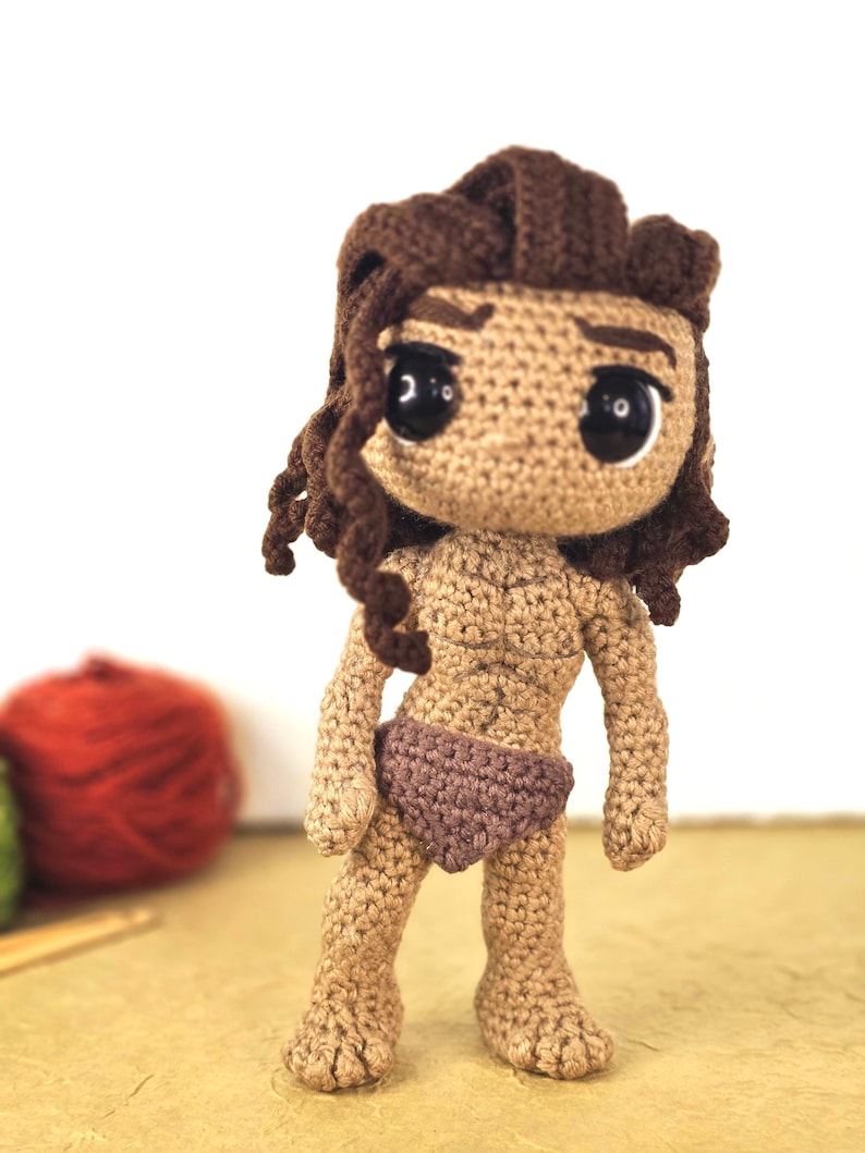 May include: A handmade crochet doll with a tan complexion, brown hair, and large black eyes. The doll is wearing a brown loincloth. The doll's hair is styled in a curly fashion. The doll is standing on a light brown surface.