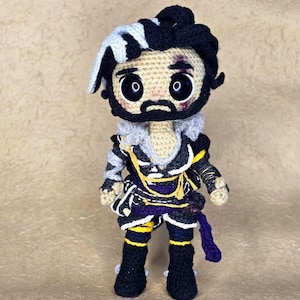 May include: A handmade crochet doll with a detailed design. The doll features a black beard, dark hair with a white streak, and a costume with purple, yellow, and black accents. The doll is standing and has large, expressive eyes.