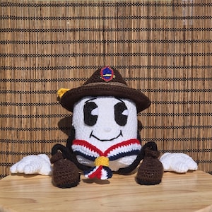 Puede incluir: Muñeca de ganchillo con una cara blanca, ojos negros y una boca sonriente. Lleva un sombrero marrón con una insignia, una cinta roja, blanca y azul y guantes marrones. La muñeca tiene alas blancas y está colocada contra una persiana de bambú.