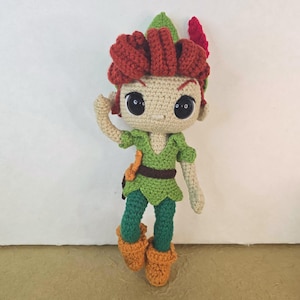 May include: A handmade crochet doll of a character with red hair, green outfit, and orange boots. The doll has large black eyes and is posed with one arm raised. The doll's outfit includes a green tunic, trousers, and a brown belt. The doll is approximately 20 cm tall.