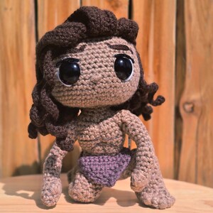 May include: A handmade crochet doll in a kneeling position. The doll has large black eyes, brown hair, and a tan skin tone. It is wearing a purple diaper. The doll is made of yarn and has a textured appearance.