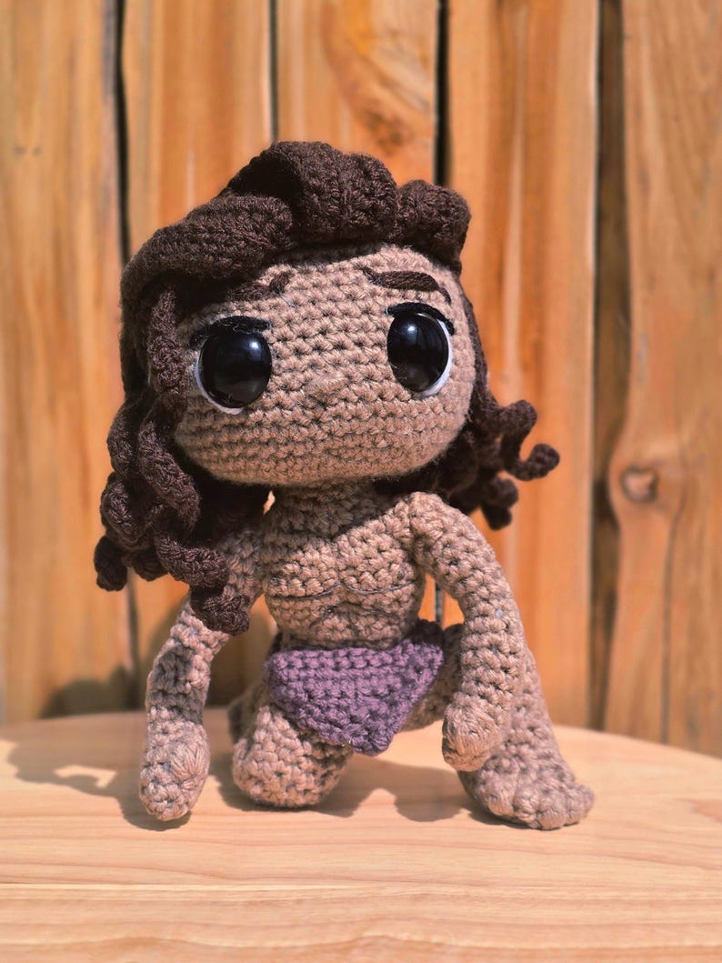 May include: A handmade crochet doll in a kneeling position. The doll has large black eyes, brown hair, and a tan skin tone. It is wearing a purple diaper. The doll is made of yarn and has a textured appearance.