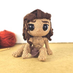 May include: A handmade crochet doll of a cartoon character, featuring a tan complexion, dark brown hair, and large black eyes. The doll is seated with its legs extended, wearing a small purple loincloth. The doll is made of yarn.